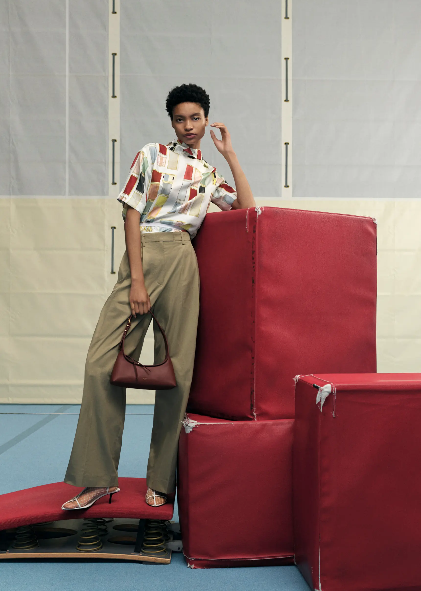 Fashion model poses in patterned shirt, wide-leg pants, and handbag leaning against a stack of red gym mats in a gymnasium.