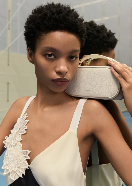 Close-up of a woman posing with a small neutral-toned purse. She wears a white dress with floral details indoors in a studio setting.