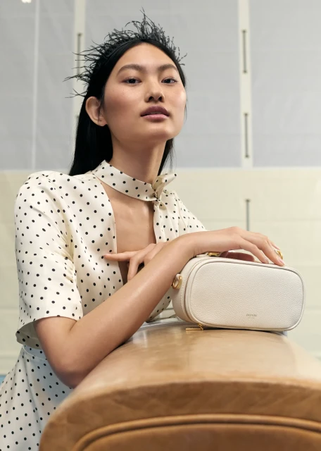 Portrait of an Asian fashion model with a cream-colored leather handbag in a photo studio with bright, soft lighting and neutral colors.