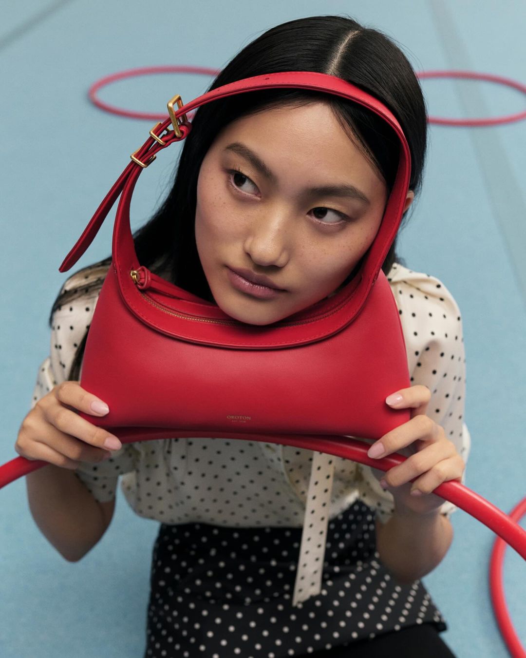 Asian model presents a red leather handbag indoors, wearing a polka dot blouse and skirt, with red hoops in the background.