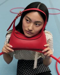 Close-up portrait of a woman holding a red leather handbag framed around her face against a pale blue background.