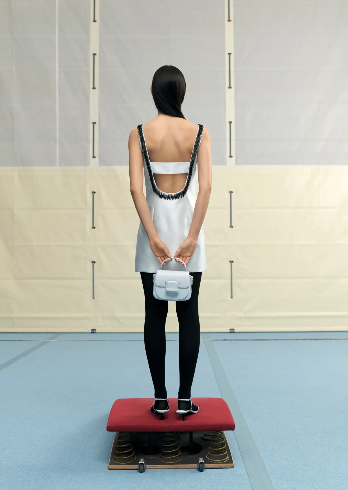 Back view of a slim woman with dark hair in a white dress, black tights, and heels, holding a light blue purse, standing indoors.