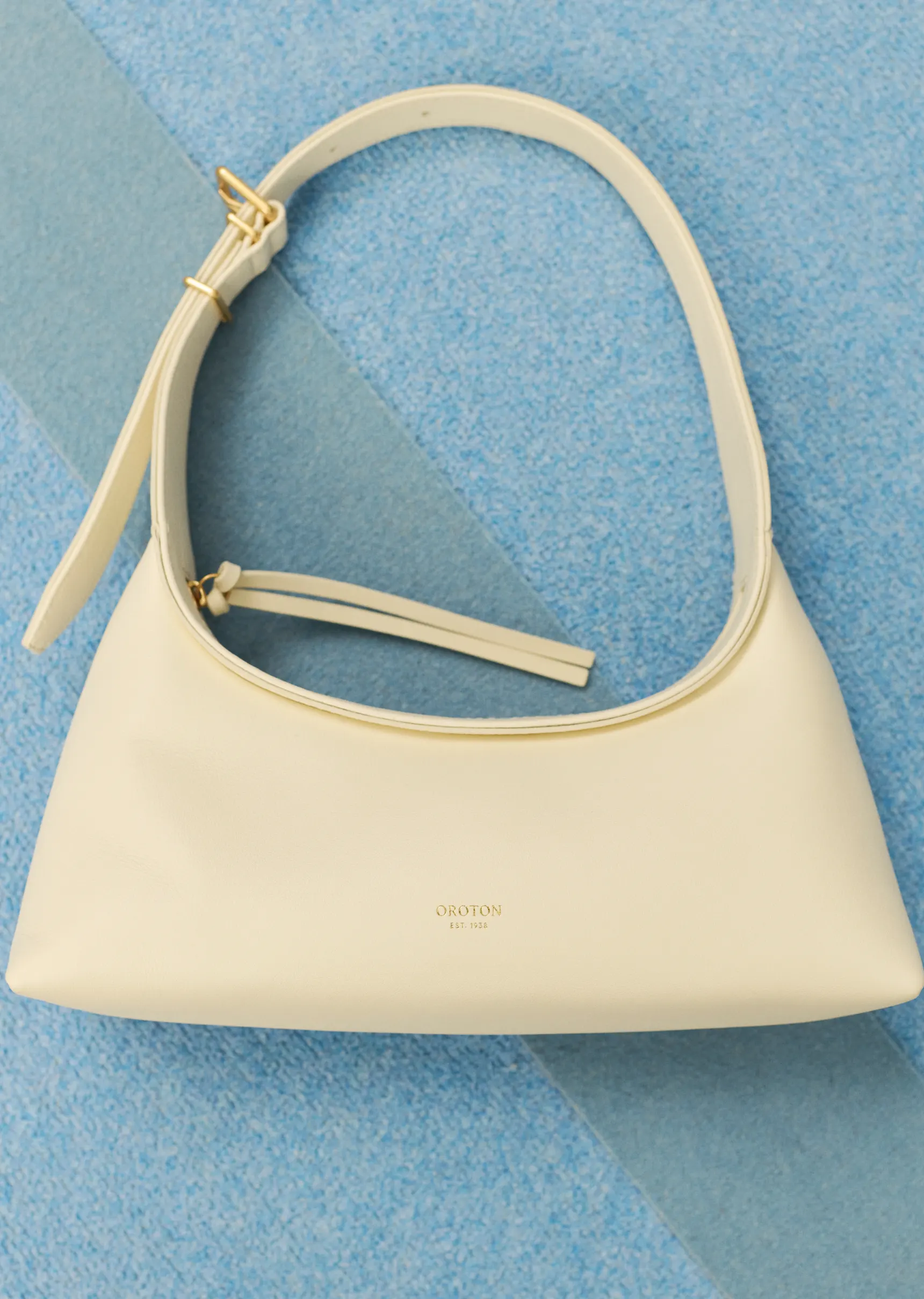 Ivory-colored handbag with a strap and metal accents, set against a textured light blue background for a clean and minimalist presentation.