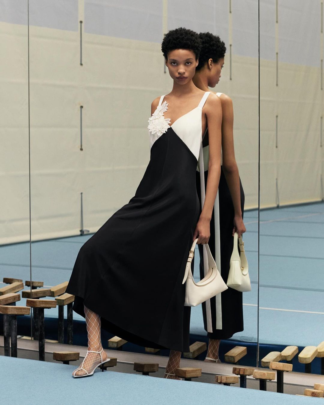 A fashion photo featuring a Black female model in a black and white dress, holding a white handbag in an indoor studio setting.