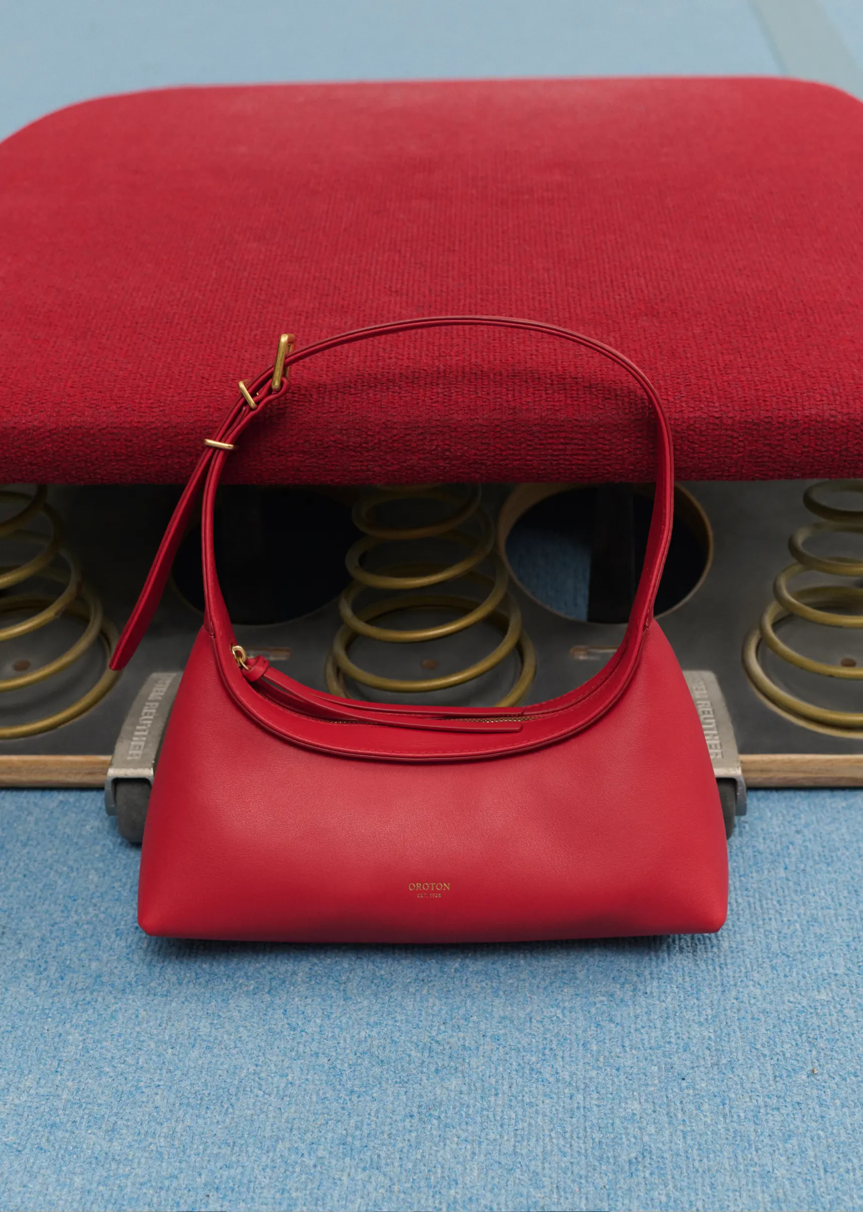 Red leather shoulder bag with gold hardware on a vintage spring stool against a pale blue background in a studio setting.