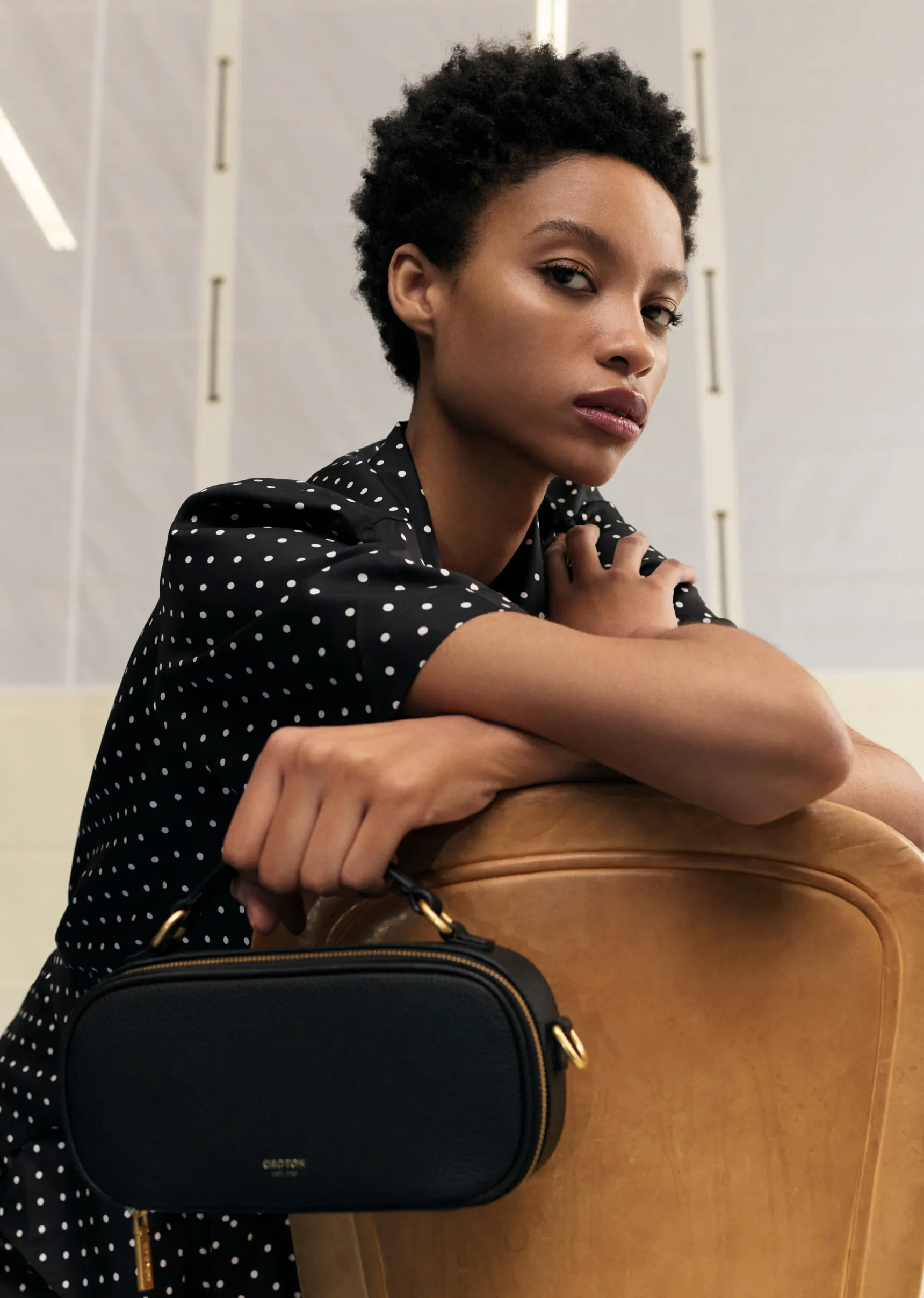 A Black woman with an afro is holding a black purse in a close up shot in a photo studio with a soft lighting.