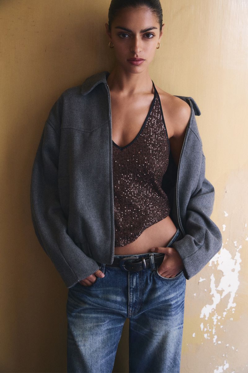 Woman in halter top and oversized jacket posing against a textured wall in a fashion editorial-style photograph with natural lighting.