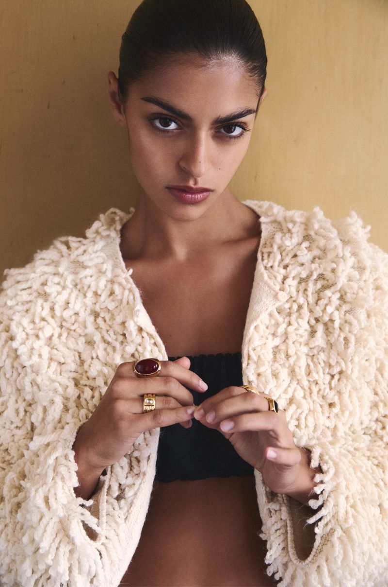 A young woman with dark hair and fair skin poses indoors wearing a fringed jacket and jewelry, looking directly at the camera.