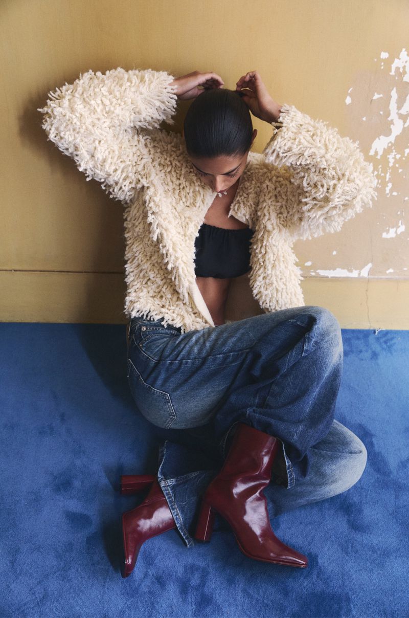 Fashion shot of a young adult woman sitting on a blue surface wearing a shaggy jacket, jeans, black top and maroon boots against a wall.