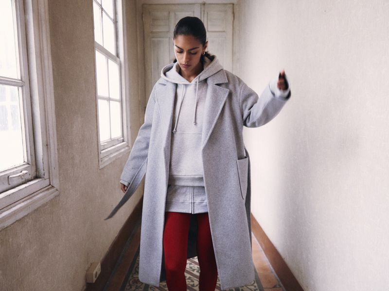 Model wearing a light grey coat over a grey hoodie and red pants walking down a hallway with windows.