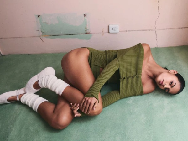 Woman in an olive bodysuit and white leg warmers poses on a green carpet, with cracked walls in the background, creating a minimalist indoor fashion portrait.