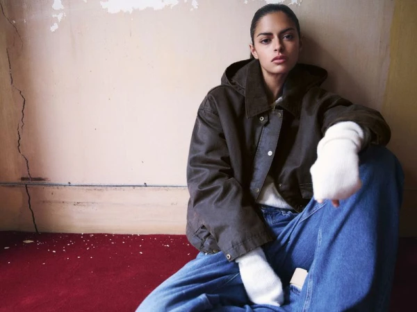 A seated woman wearing a dark brown hooded jacket and blue jeans against a textured wall and red floor.