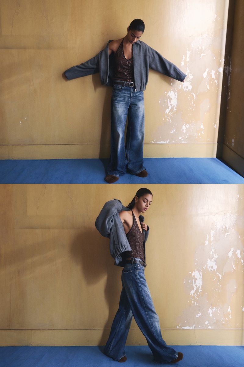 A woman models wide-leg jeans and a short jacket in a photo studio with a blue floor and textured wall in a fashion editorial shot.