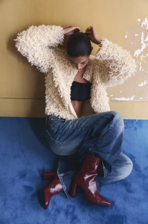 Model seated on a blue carpet against a wall, wearing a textured cream jacket, black top, denim jeans, and red boots.