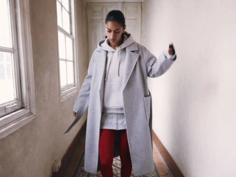 Model wearing a light grey coat over a grey hoodie and red pants walking down a hallway with windows.