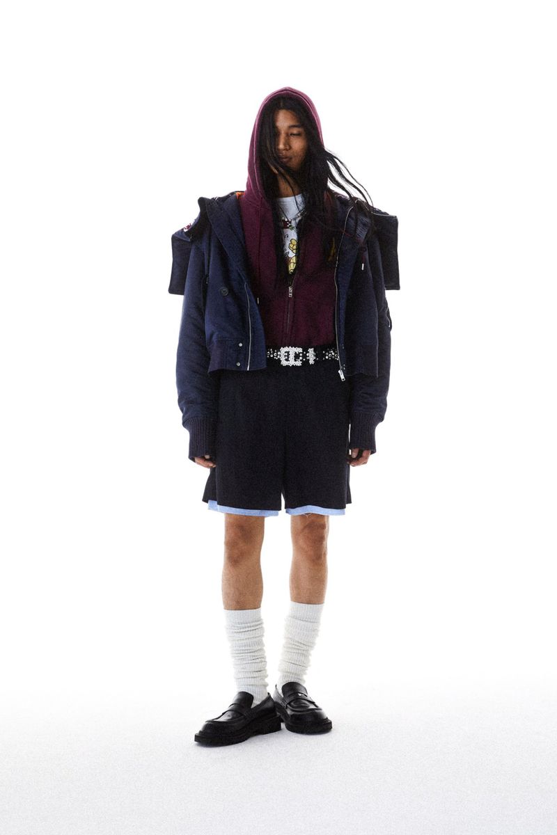 Model wearing a navy cropped jacket, burgundy hoodie, black layered shorts, slouchy white socks, and loafers against a white backdrop.