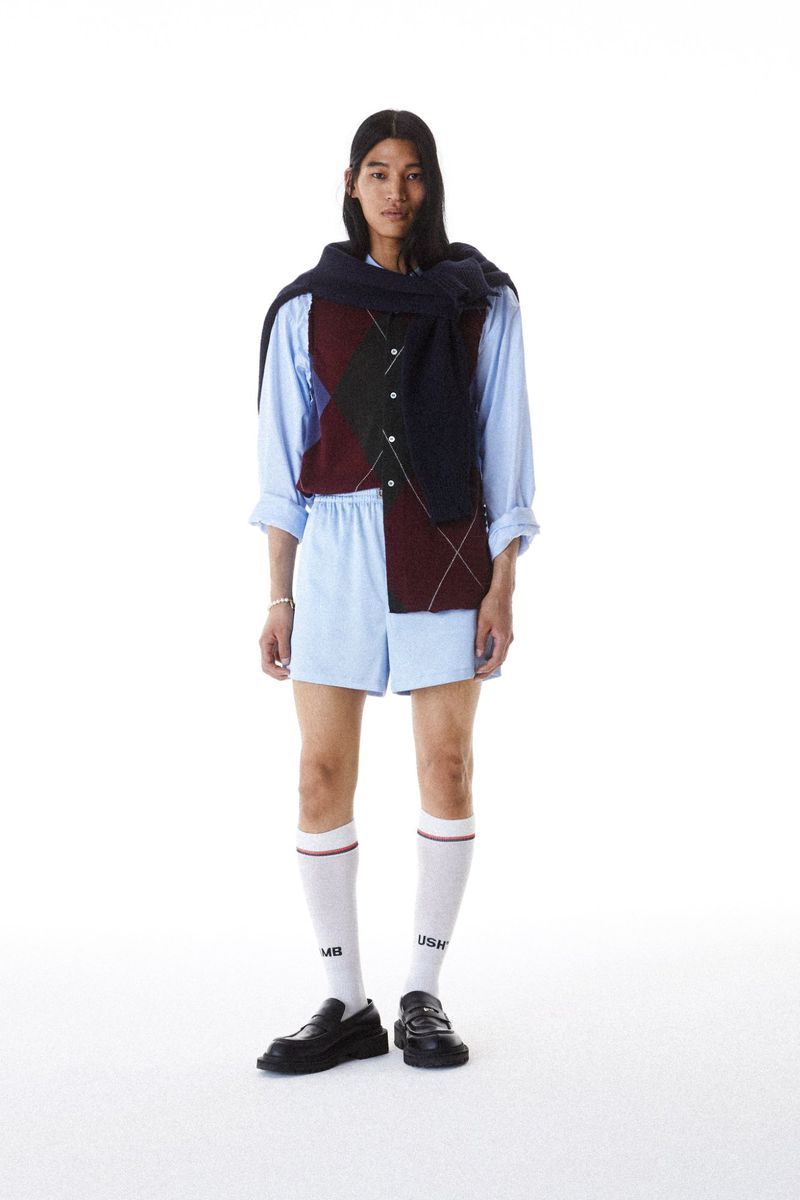 Full-length studio shot of an adult male in an argyle vest, blue shirt, shorts, knee socks, and loafers against a white backdrop. 