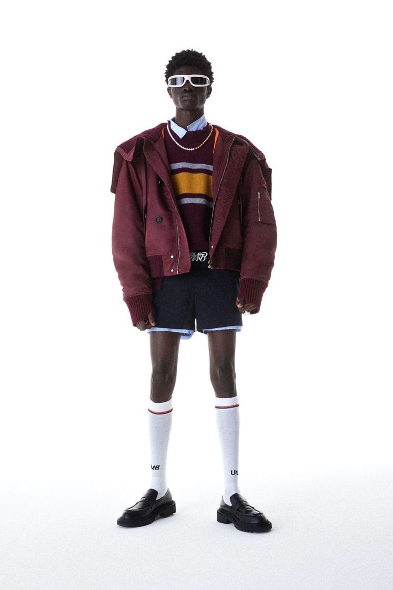 Dark-skinned male fashion model in a burgundy jacket, striped sweater, and shorts, posing against a white backdrop in a full shot.