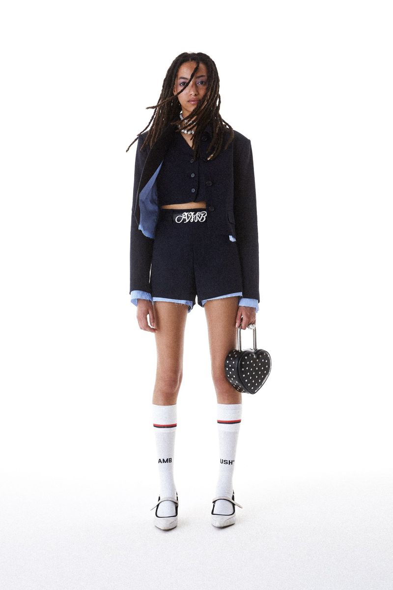 A young woman with dreadlocks poses in a cropped blazer and shorts, holding a heart-shaped purse against a white backdrop.