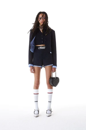 A young woman with dreadlocks poses in a cropped blazer and shorts, holding a heart-shaped purse against a white backdrop.