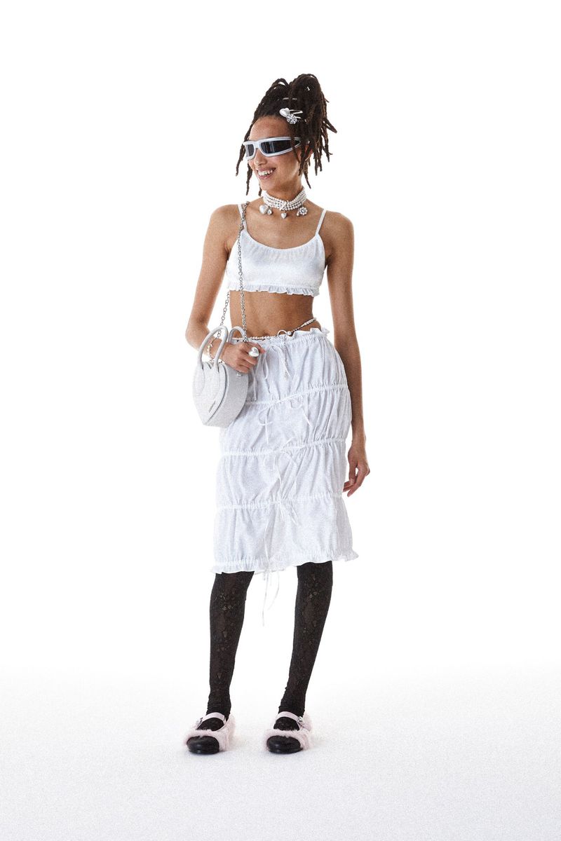 A full-shot of a smiling woman with dreadlocks wearing a white crop top and skirt with black stockings and sandals against a white backdrop.