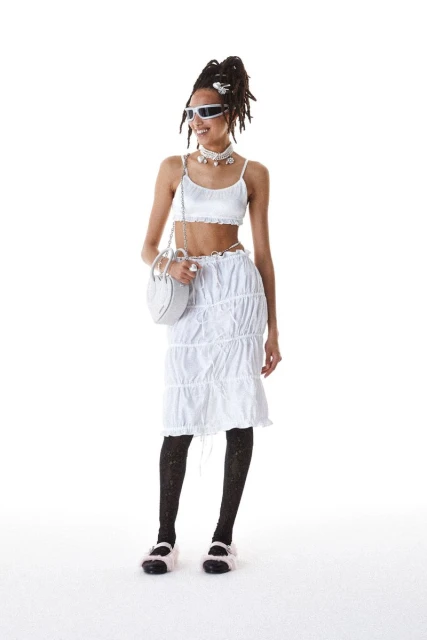 A full-shot of a smiling woman with dreadlocks wearing a white crop top and skirt with black stockings and sandals against a white backdrop.
