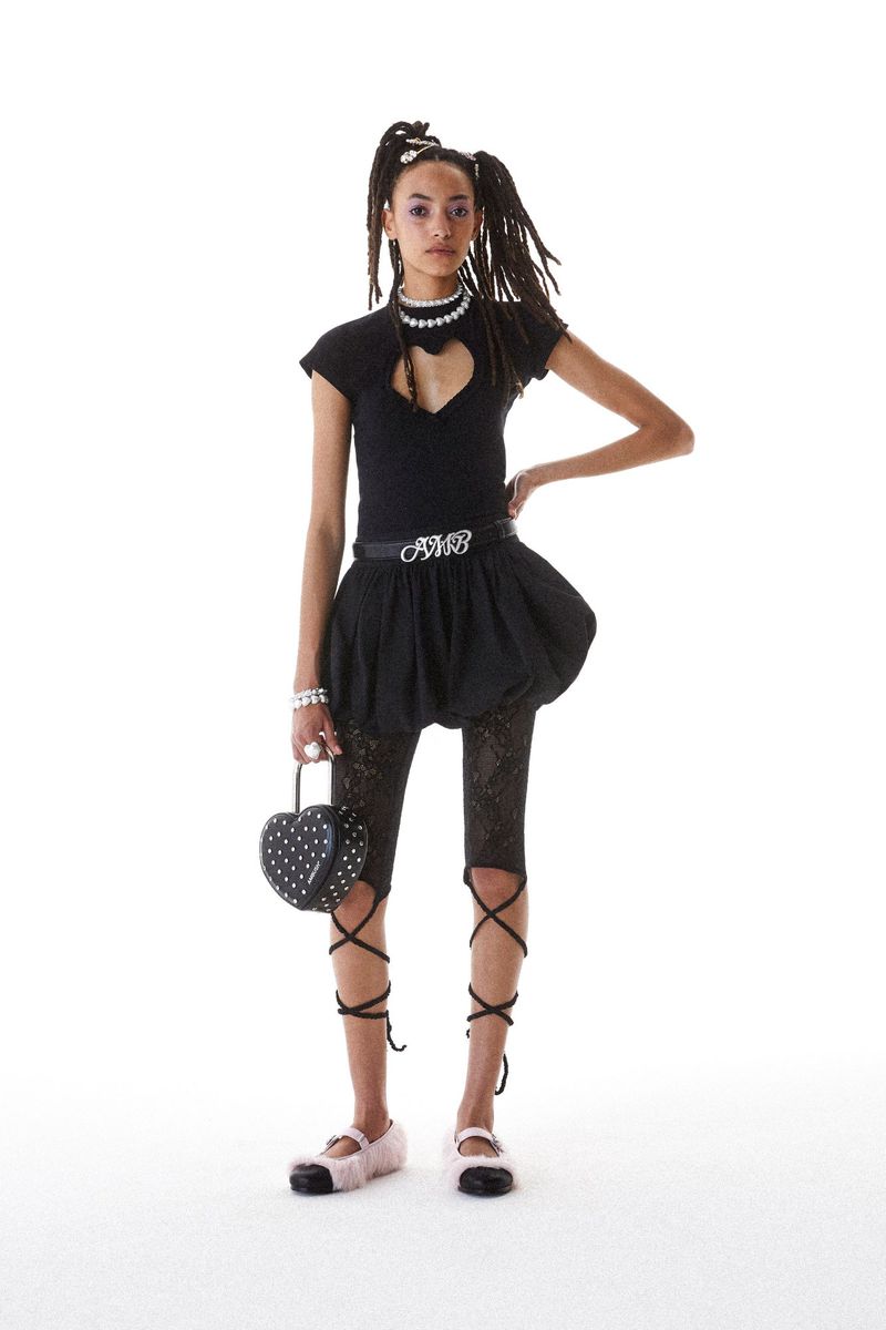 Fashionable young adult woman strikes a pose in a studio, sporting a black top with a heart cutout, a bubble skirt, and lace-up leg coverings.