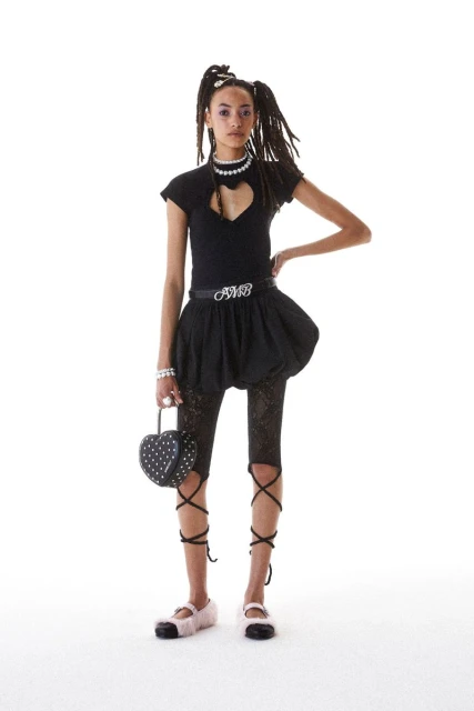 Fashionable young adult woman strikes a pose in a studio, sporting a black top with a heart cutout, a bubble skirt, and lace-up leg coverings.