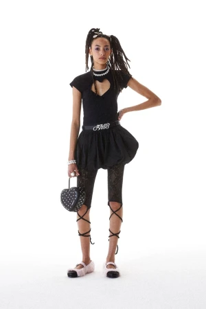 Fashionable young adult woman strikes a pose in a studio, sporting a black top with a heart cutout, a bubble skirt, and lace-up leg coverings.