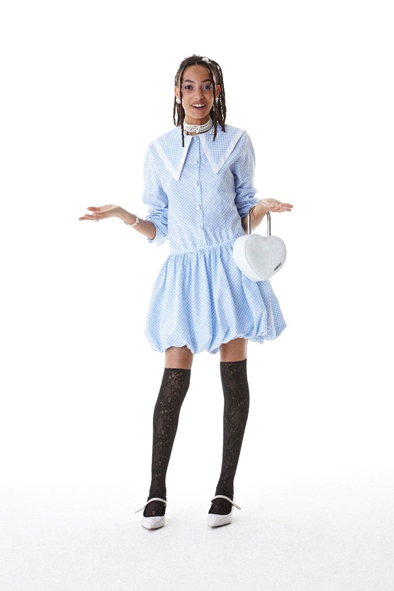Full-shot of a young adult woman wearing a light blue polka-dot dress and black patterned stockings, holding a heart-shaped white purse.