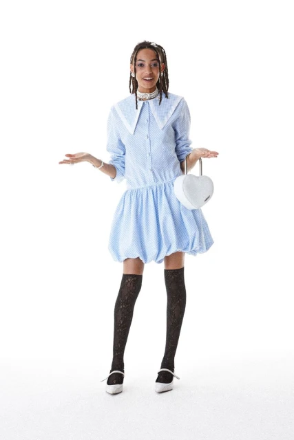 Full-shot of a young adult woman wearing a light blue polka-dot dress and black patterned stockings, holding a heart-shaped white purse.