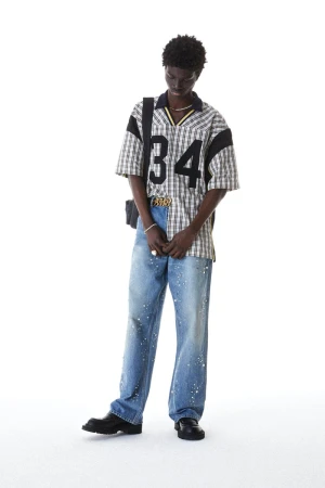 A slender adult male poses in a cream and black plaid shirt with the number 34 on the front, paired with light blue paint-splattered jeans.