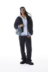Model posing in futuristic sunglasses, a dark zip jacket, light blue polo, and relaxed trousers for the Ambush S/S 25 lookbook.