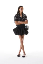 Model standing with crossed arms wearing a dark patterned shirt, flared skirt, and heart-shaped handbag against a white background