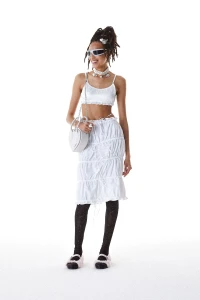 Model wearing a white crop top and ruched skirt with futuristic sunglasses and black tights against a white seamless background.