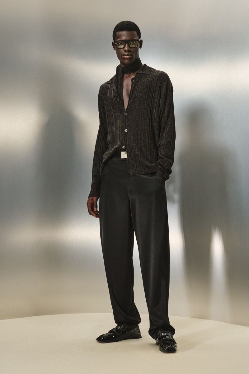 A stylish black man poses in a relaxed stance wearing a dark brown button-down shirt, black slacks, black glasses and black loafers in studio.