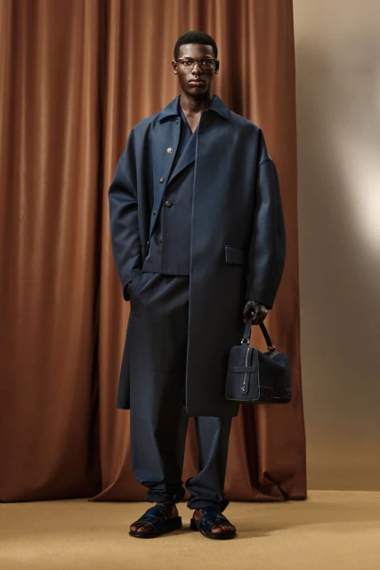 A dark-skinned man wearing a blue outfit, sandals and holding a dark blue leather bag stands in front of a brown curtain.