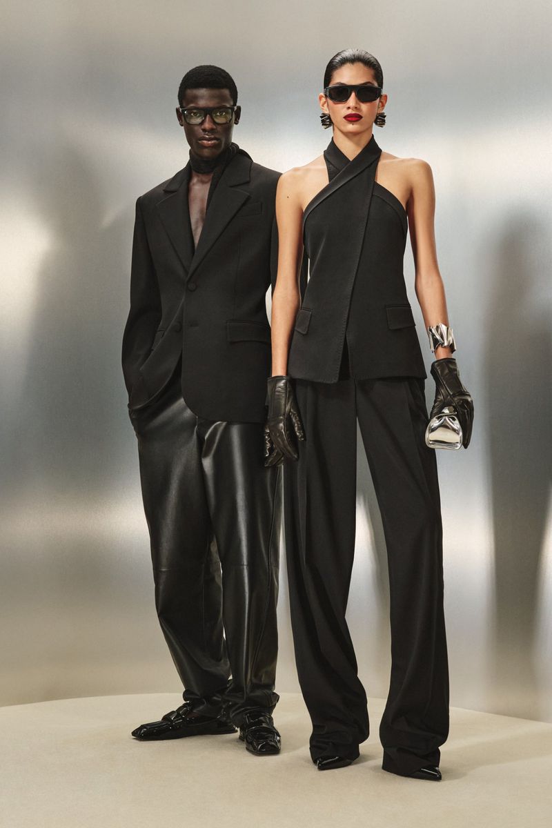 A Black man and woman model black fashion, with the man in a suit and the woman in an halter top and pants, against a silver backdrop.