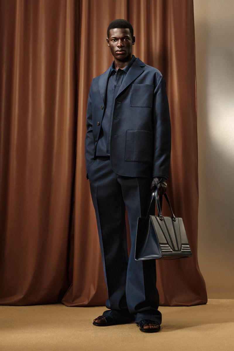 Elegant Black man in a navy suit and sandals holding a structured tote bag, standing indoors against a backdrop of brown drapery.