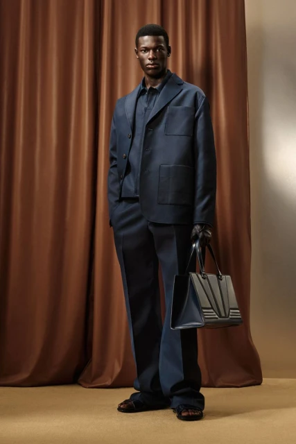 Elegant Black man in a navy suit and sandals holding a structured tote bag, standing indoors against a backdrop of brown drapery.