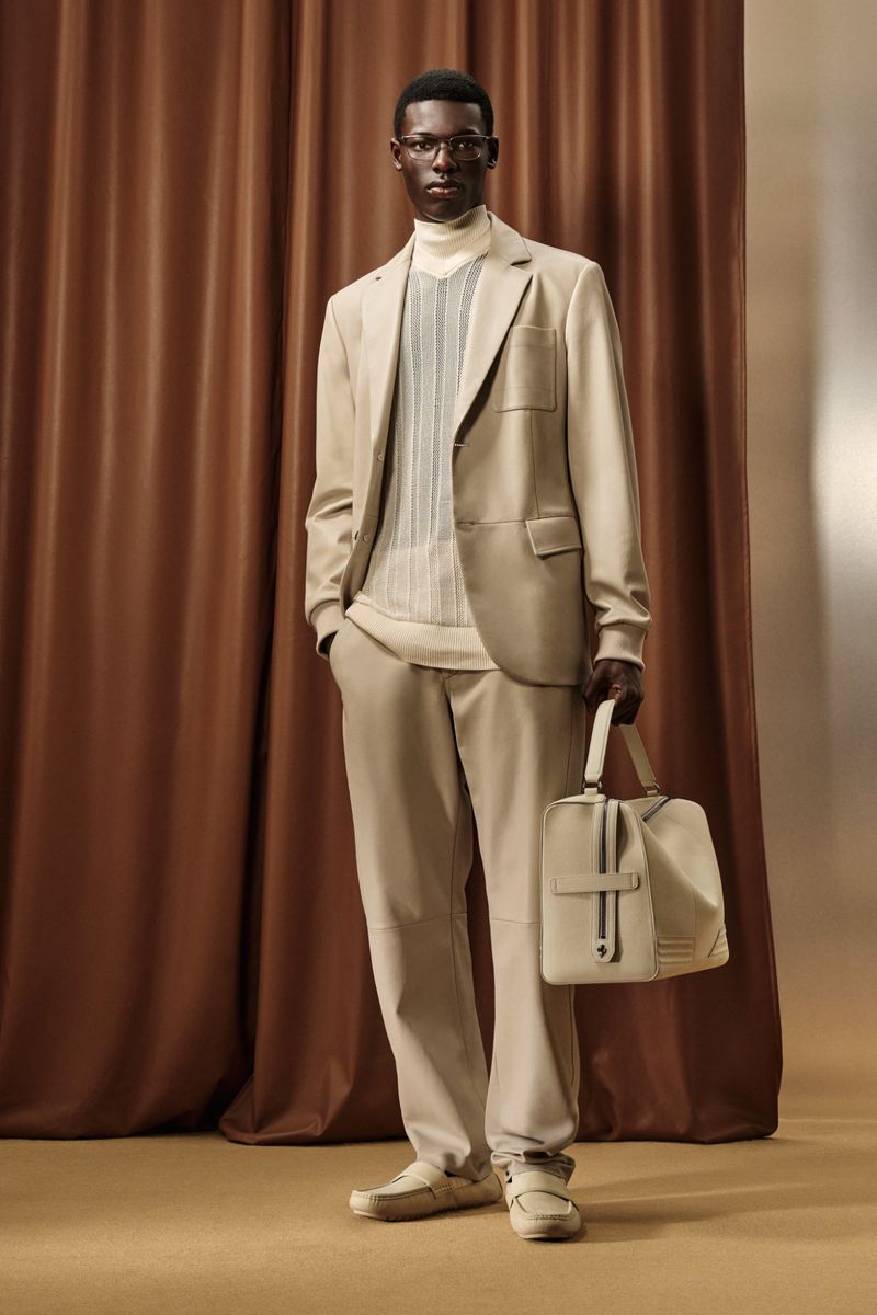 Man in tan suit and glasses stands in front of brown curtain, holding tan bag. Fashion editorial photography in warm neutral tones.