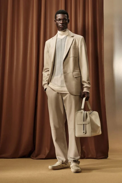 Man in tan suit and glasses stands in front of brown curtain, holding tan bag. Fashion editorial photography in warm neutral tones.