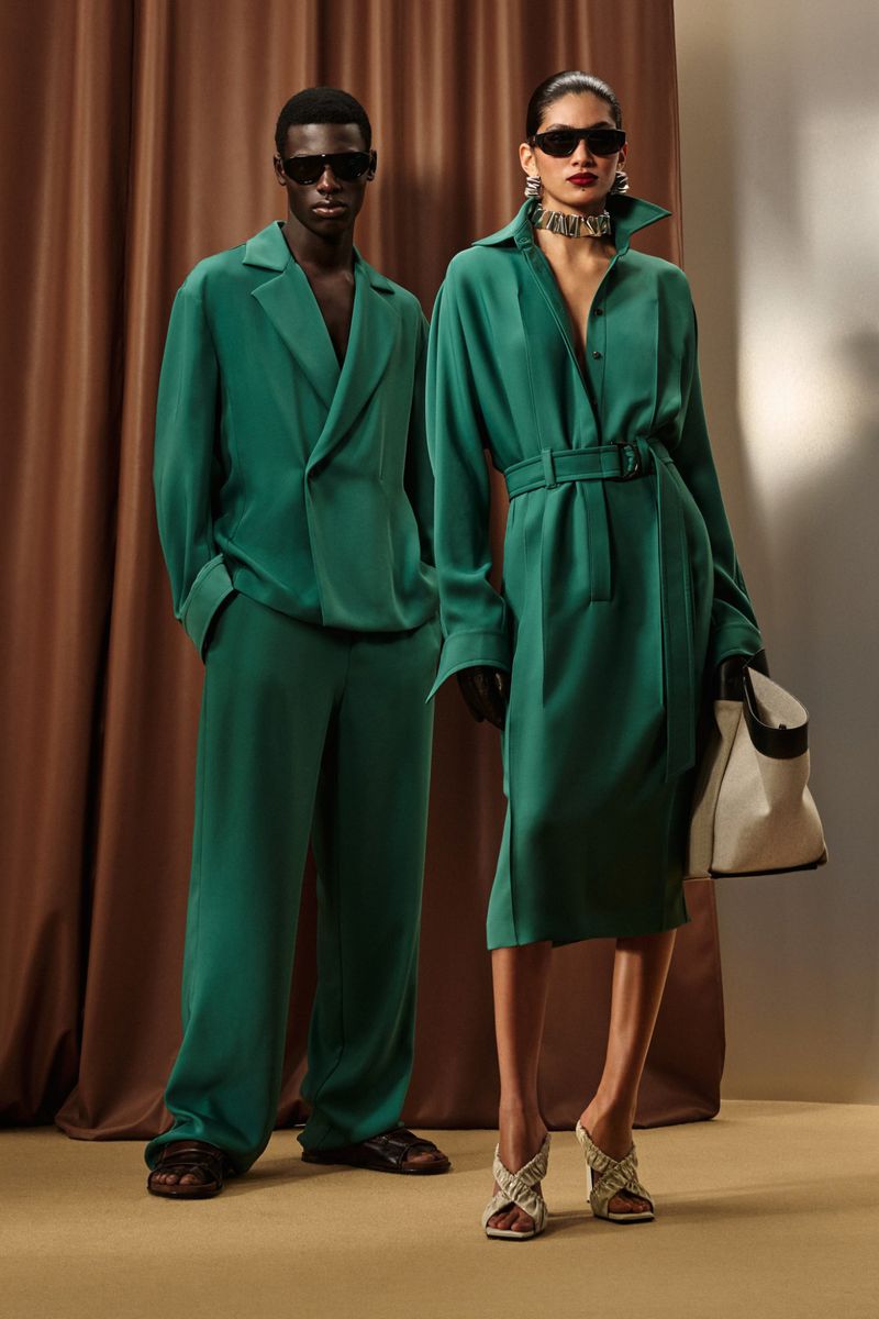 Two fashion models pose indoors against a brown curtain, wearing matching green outfits and dark sunglasses, holding a tote bag.