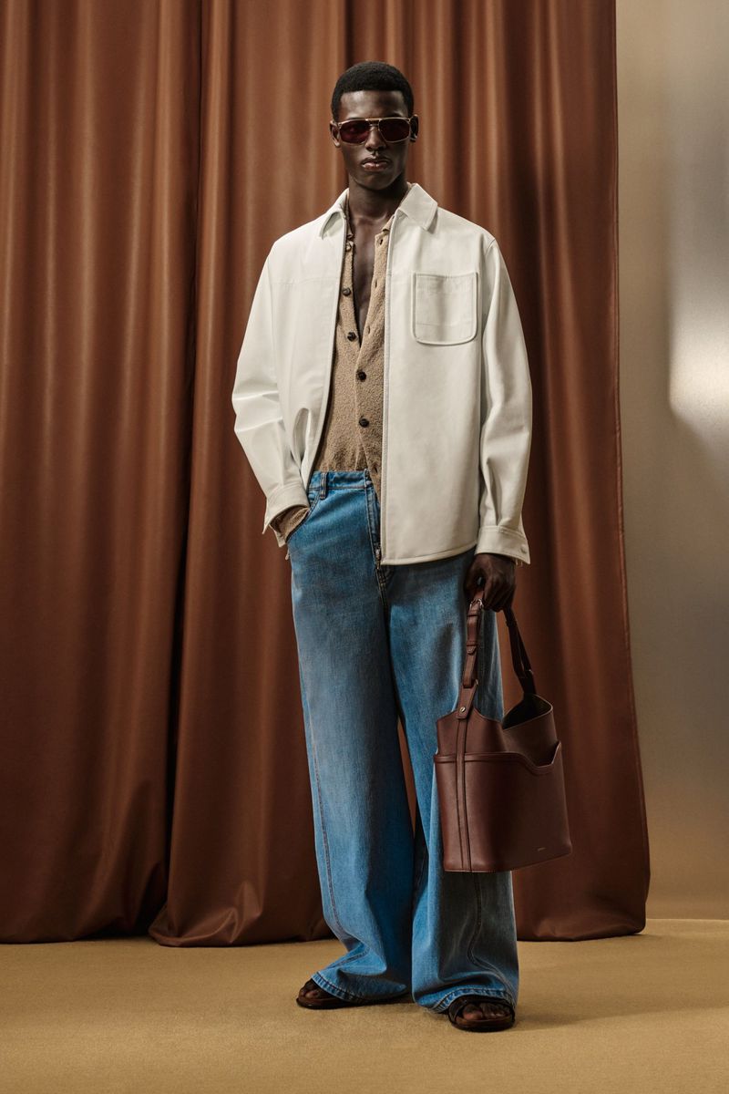 Fashionable black male model posing in stylish denim jeans, open-front shirt, and brown handbag against a brown curtain backdrop.