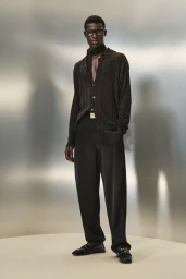 Model Victor Shalomi wears a black knit shirt, trousers, neck scarf, and glasses against a metallic backdrop for Ferrari Resort 2025.