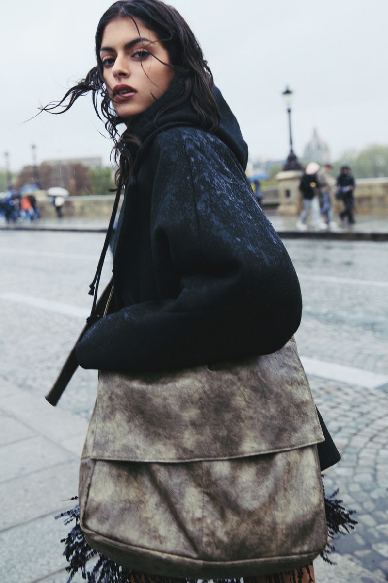 Woman in hoodie and large fringe bag walks on urban street in outdoor setting. Pedestrians and historic architecture appear in the background.