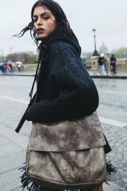 Woman in hoodie and large fringe bag walks on urban street in outdoor setting. Pedestrians and historic architecture appear in the background.
