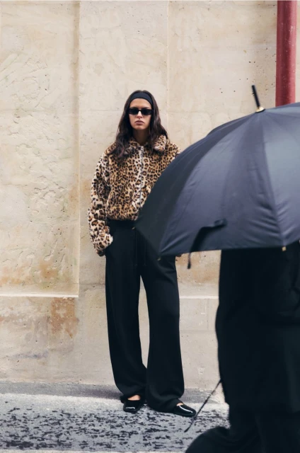 A woman with long brunette hair and sunglasses wears a leopard-print jacket and stands against a wall with an umbrella in the foreground.