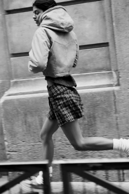 Black and white image of a woman running in a plaid skirt and hooded jacket in an urban setting, captured with a blurred motion effect.