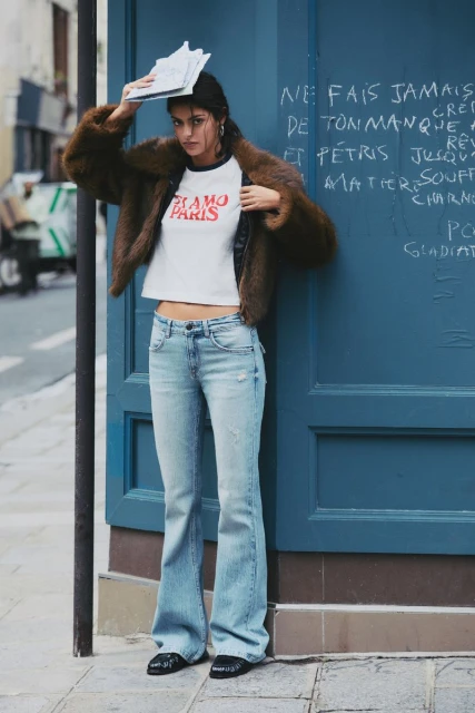Woman in brown fur coat and jeans leaning against blue door, holding papers overhead, in urban setting. Fashion editorial style.